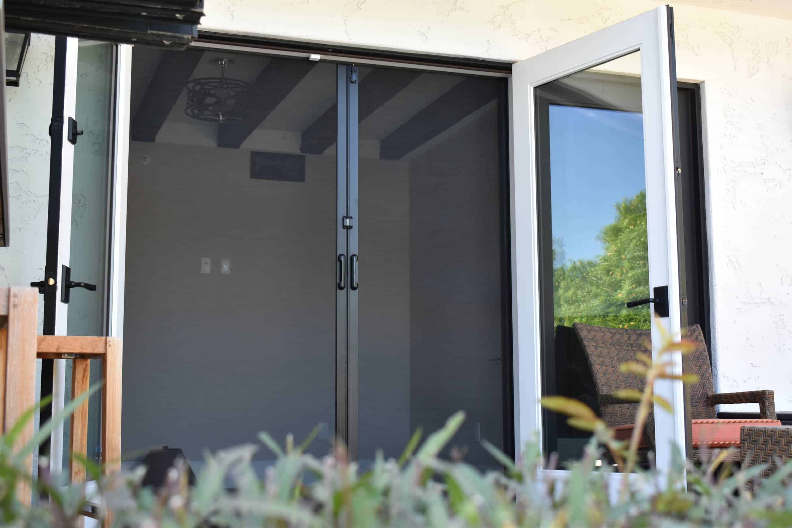 Retractable screen doors installed on a patio door, allowing fresh air while keeping insects out. The black mesh blends seamlessly with the open outdoor space.